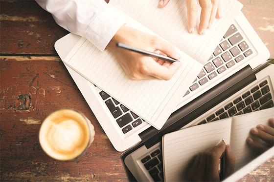 woman writing in notebook on top of laptop